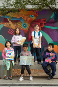 A group of kindergarten and first-grade students pose with their favorite books in front of a school mural.